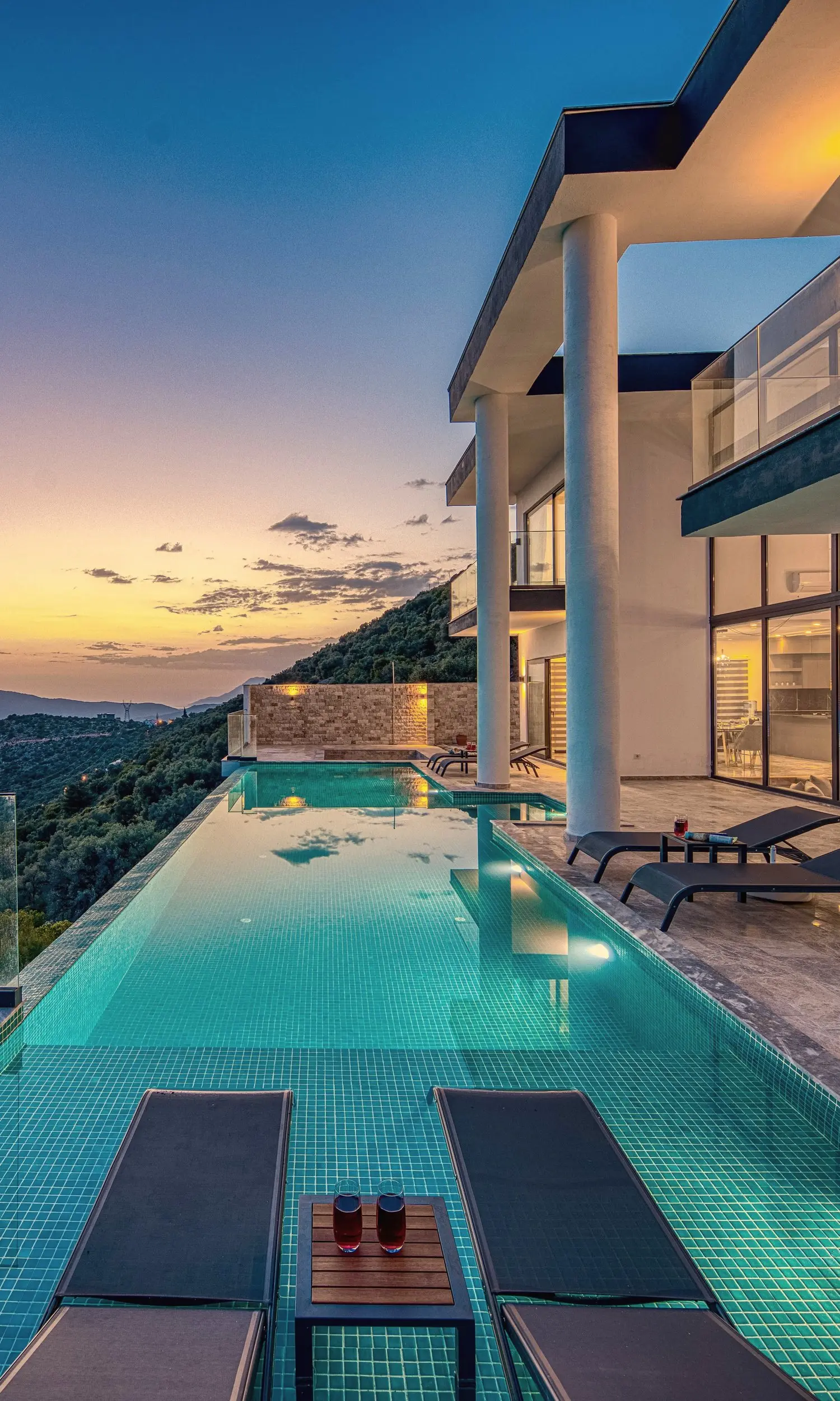 A contemporary villa of white columns, glass and travertine terraces at dusk, its infinity pool running out towards wooded hills and the sea.